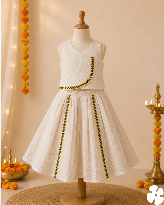 A sleeveless white textured dress displayed on a wooden mannequin, decorated with green and yellow braided trim details. The background is a warm beige decorated for a festive occasion with marigold garlands, a diya, and a bowl of marigold flowers.